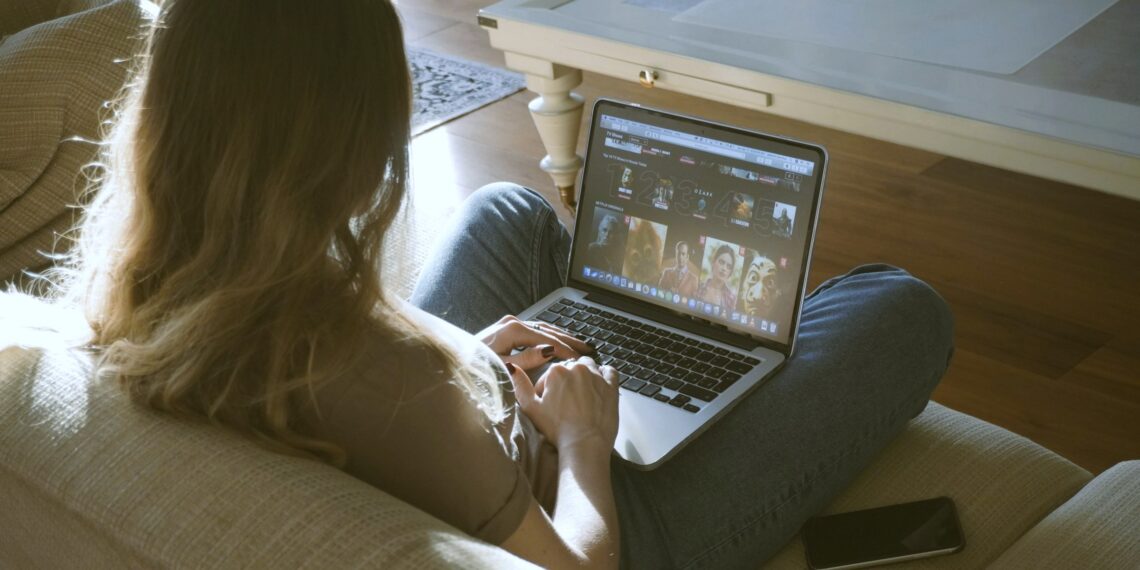 a woman sitting on the couch browsing movies on her laptop